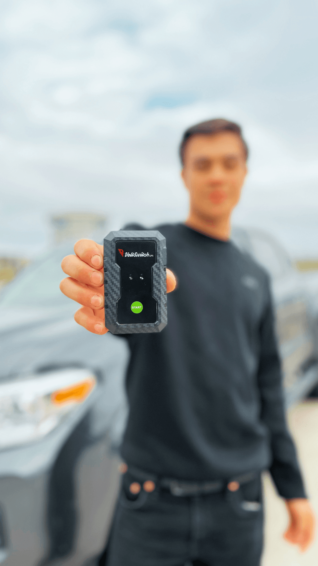 Hand holding a black VoltSwitch GPS tracker with a green start button, blurred car background.