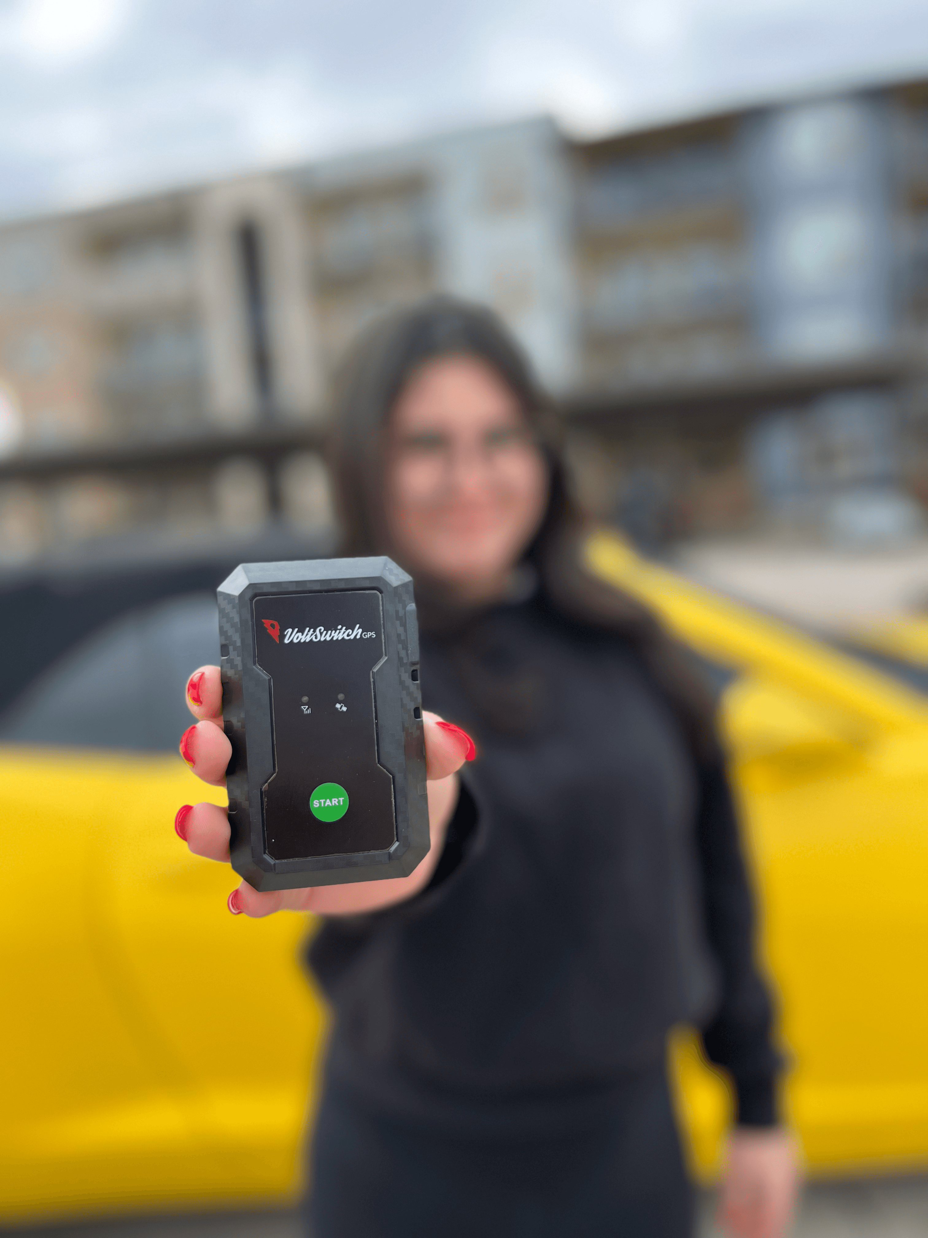 A woman displays a black VoltSwitch GPS tracking device in front of a bright yellow car.
